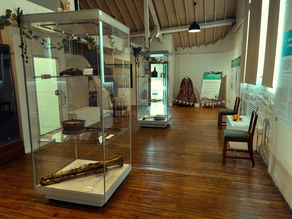This image depicts an indoor view of a museum display. The room has wooden flooring and a vaulted ceiling with visible beams. The centerpiece is a glass display case with a brass ophicleide instrument lying on a white cloth at its base. Surrounding items include a small drum and a violin. The case reflects the surrounding room, slightly obscuring the view of another exhibit behind it, featuring a tepee-like structure adorned with a floral wreath. To the right, informational panels can be seen, detailing aspects of the museum's exhibits. The scene is softly lit by overhead lighting, creating a warm ambiance.