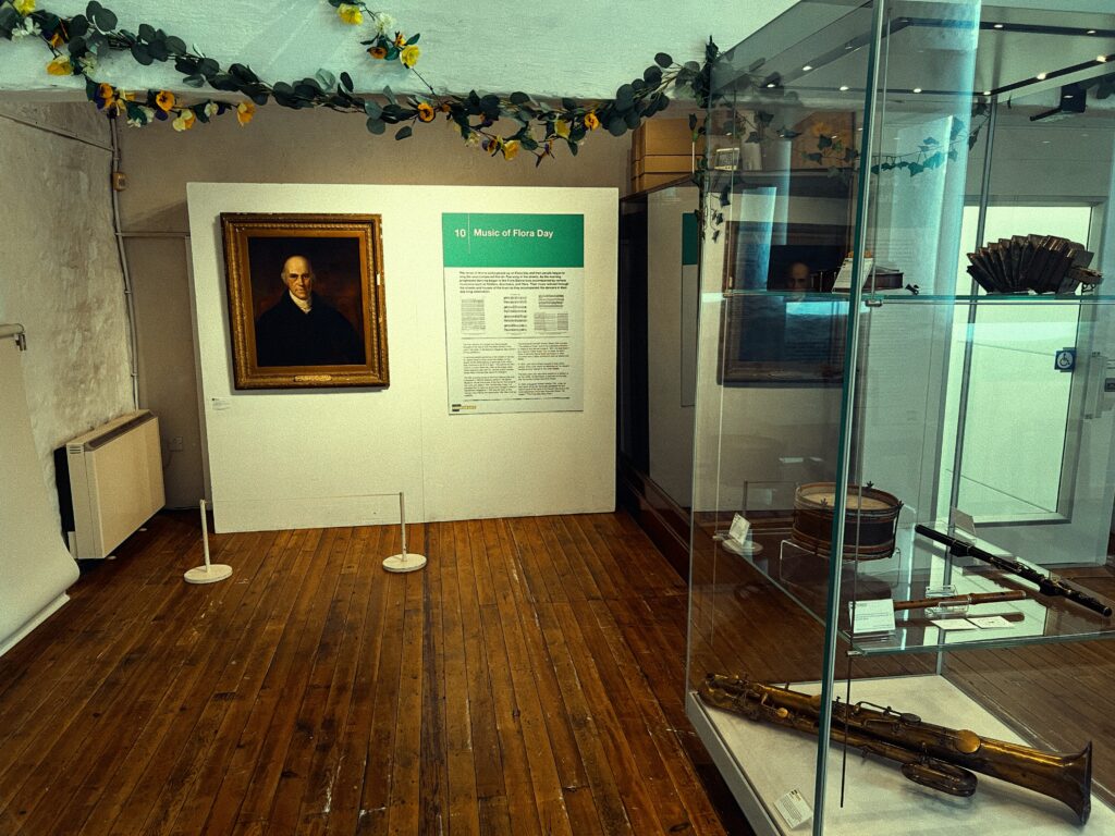 A different part of the exhibition, showing a portrait painting on the left wall, which depicts a solemn-faced individual, Davies Gilbert FSA, in formal attire. To the right, another glass display case visible from the first image holds the brass ophicleide, along with a drum, pipes, and a black concertina. A green informational panel adjacent to the case discusses the music of Flora Day. The museum's wooden flooring, soft lighting, and a part of the ceiling structure contribute to the setting's historical ambiance.