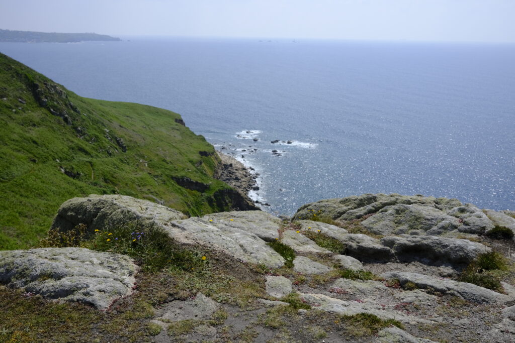 This photo depicts a stunning coastal scene with a focus on a rocky cliffside overlooking the sea. In the foreground, large, weathered rocks are interspersed with patches of grass and small wildflowers, adding a touch of colour to the rugged terrain. The cliffside slopes down to meet the ocean, with a mix of green vegetation and exposed rock formations. The water below is a deep blue, shimmering in the sunlight, and stretches out to the horizon. Small waves break against the rocky shoreline. In the distance, the coastline continues, fading into a hazy outline. The sky is clear with a soft blue hue, enhancing the overall serene and expansive atmosphere of the coastal landscape.