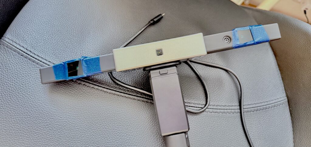 The photo shows a handheld structured light 3D scanner, laid on a leather surface. The device has a central rectangular body with sensors and cameras extending on either side. To make it more usable outdoors, temporary “shades” have been improvised: pieces of blue tape hold coverings over the infrared sensors at each end, helping reduce interference from bright sunlight. This kind of adaptation allows the scanner, which normally works best indoors or in controlled lighting, to capture reliable 3D surface data in outdoor heritage contexts.