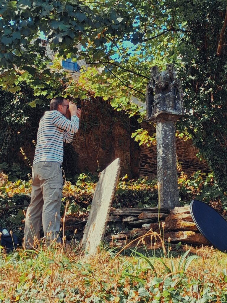 The photo shows a person in a striped long-sleeve shirt and light trousers photographing a medieval stone monument outdoors. The monument stands on a tall, weathered shaft with a carved top that looks like a lantern cross or a wayside shrine, decorated with figures. The setting is leafy and shaded, with ivy-covered walls and overgrown vegetation, suggesting a historic churchyard or village site. Nearby on the ground are equipment pieces, including a reflector disc, used to control lighting for photography. The scene captures the act of carefully documenting heritage in a tranquil, historic environment.
