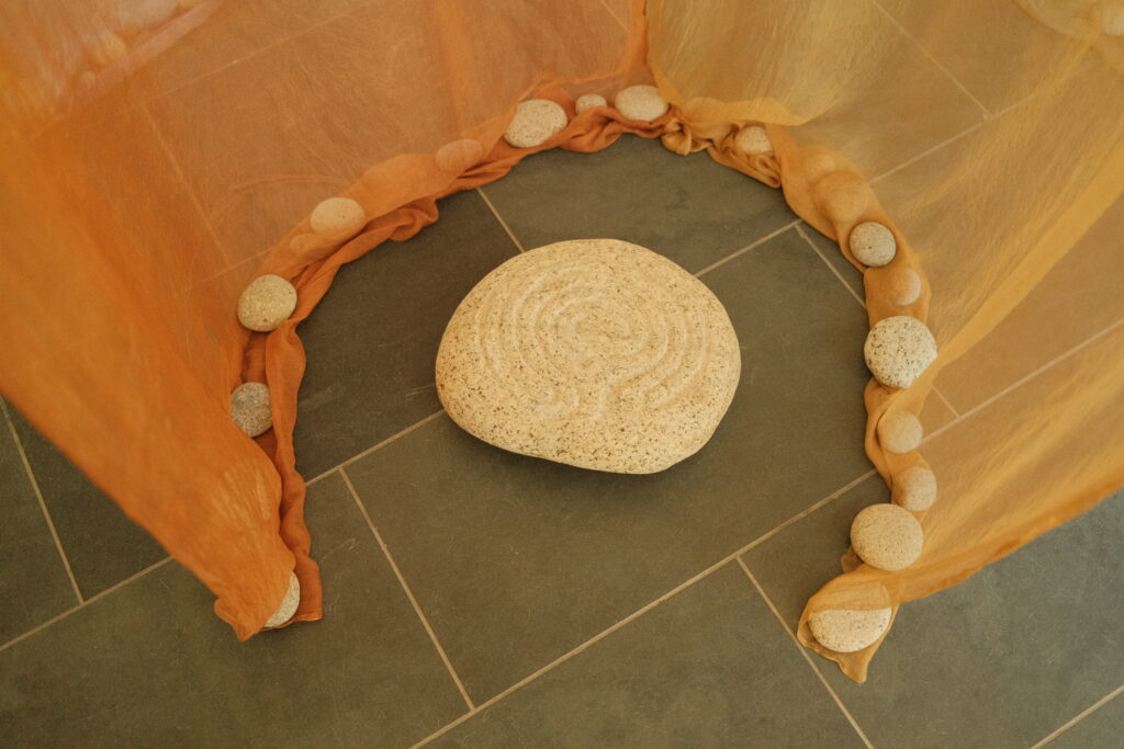 A circular stone with a carved labyrinth pattern placed on a tiled floor, framed by an orange fabric draped on the ground and held in place with smaller weather beach stones.