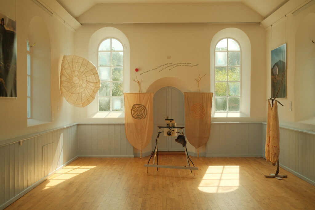 A bright gallery room with tall arched windows, wooden floor, and fabric banners with spiral designs hanging by a central door. Sculptural objects and textiles are arranged around the space, with soft sunlight streaming through the windows.