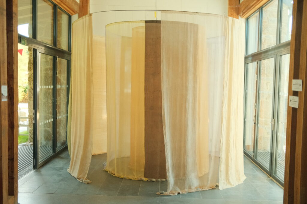 A circular art installation made of tall, sheer fabric panels in soft beige tones, hanging from a frame, column, placed inside a bright room with glass walls and wooden beams.