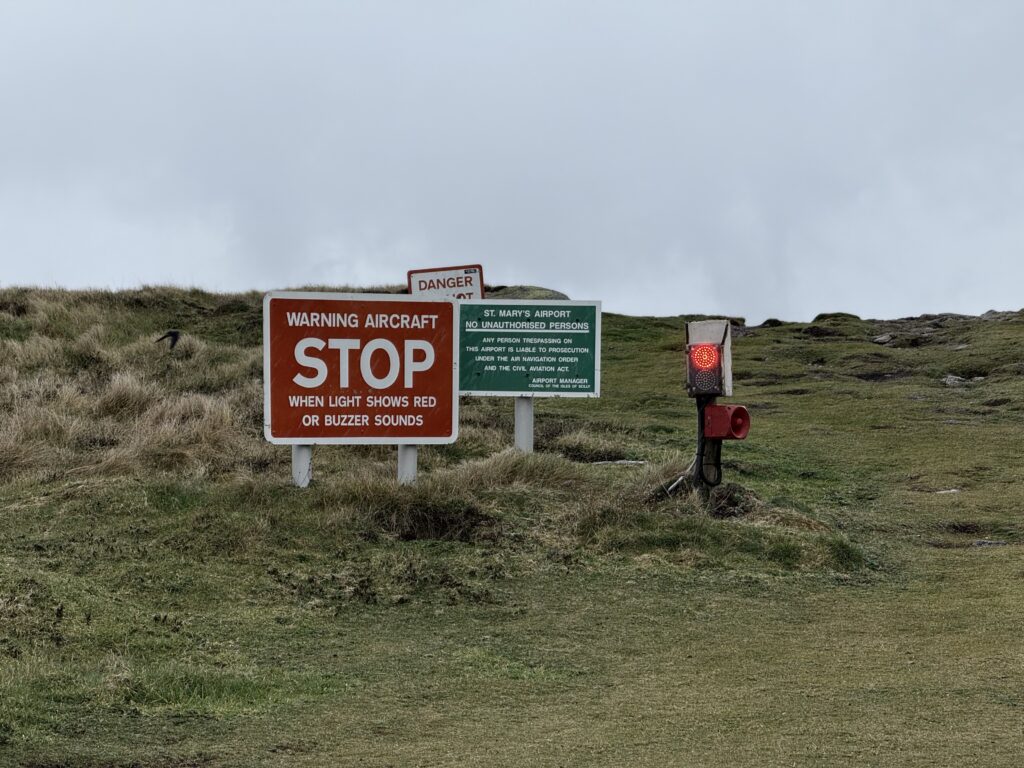 A grassy clifftop showing a large red sign reading "Warning Aircraft - STOP - when light shows red or buzzer sounds" next to a green sign stating that the land belongs to St Mary's Airport. Next to that is an illuminated red light with the buzzer fixed below it.
