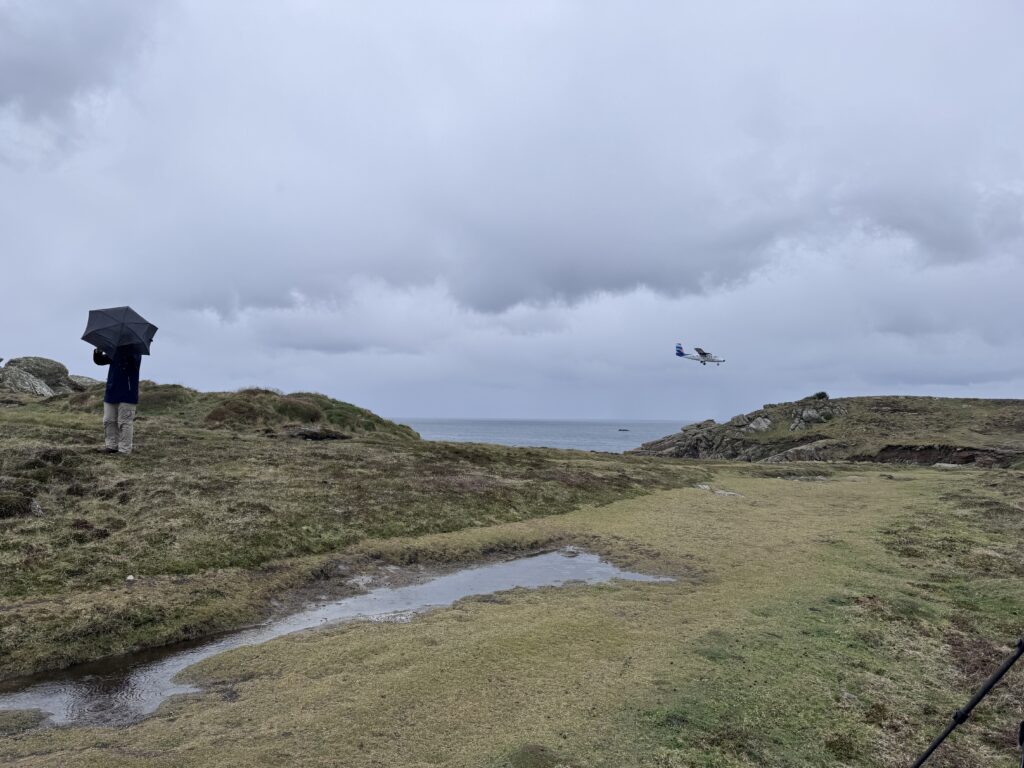 A coastal scene with granite outcrops, grassland and sea visible. To the left is a man with an umbrella. To the right is a small aeroplane coming in to land on the distant runway.